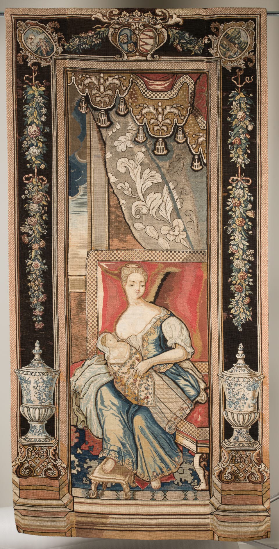 Bild av en broderad tapet med motiv av en kvinna med ett barn i famnen sittande på en röd stol, omgiven av blomrankor.