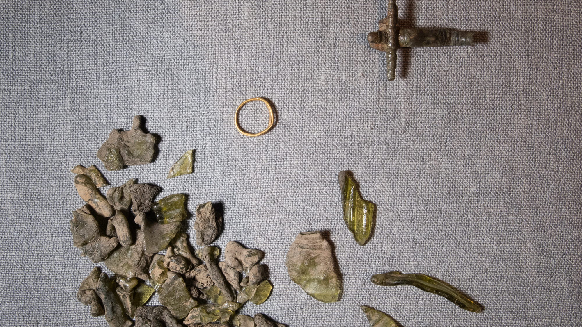 Olika fynd som hittats vid arkeologisk utgrävning, bland annat en ring, ihopsmält glasfragment och ett kors.