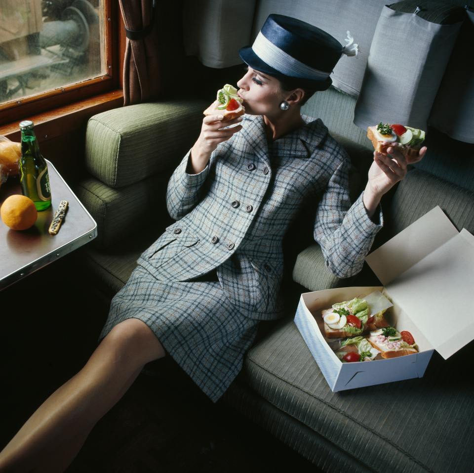 Kvinna i dräkt och hatt sitter i en tågkupé och äter smörgåsar ur en medhavd kartong 