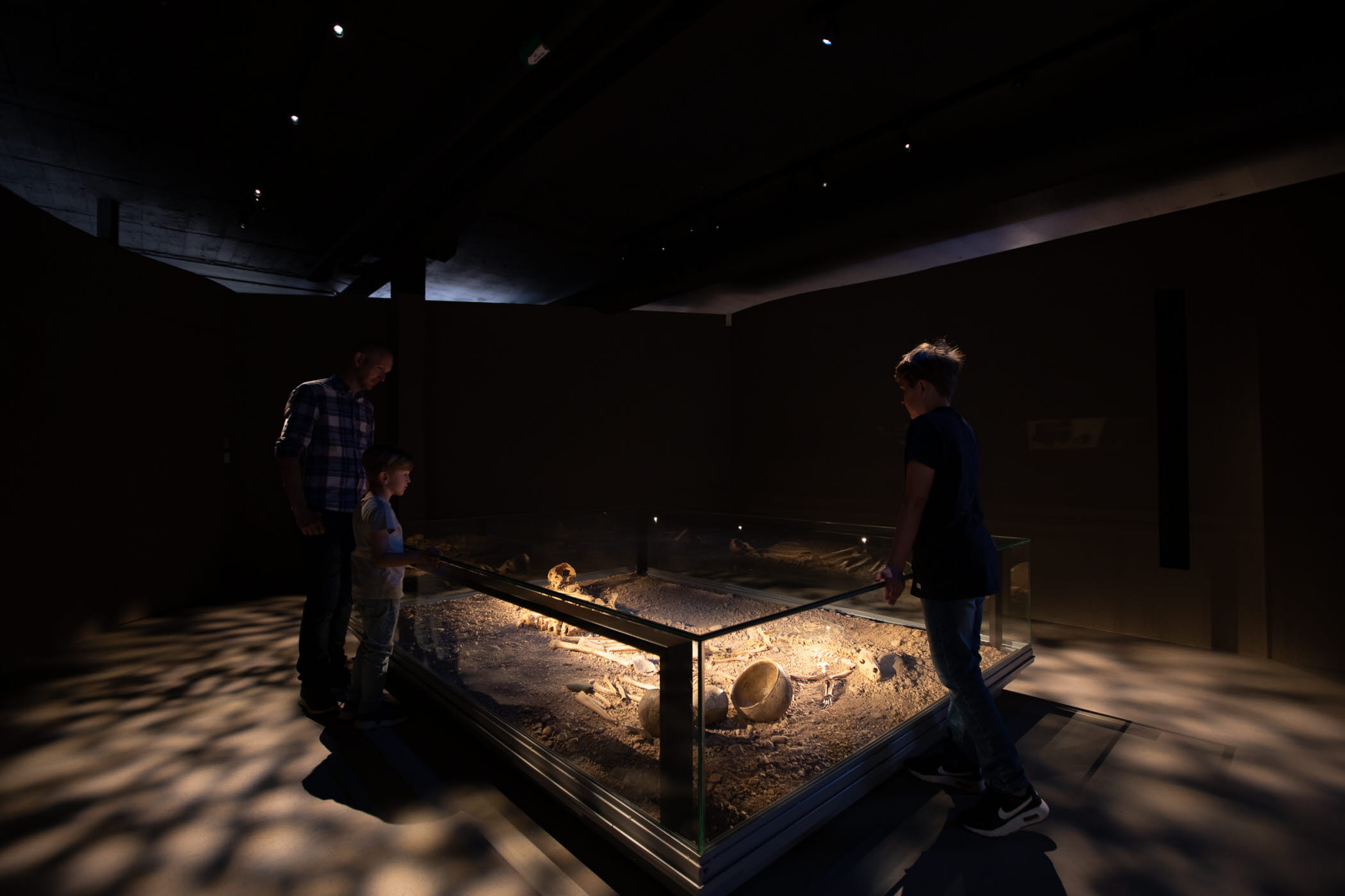 Två barn och en vuxen står i ett mörkt rum runt en upplyst inglasad grav.