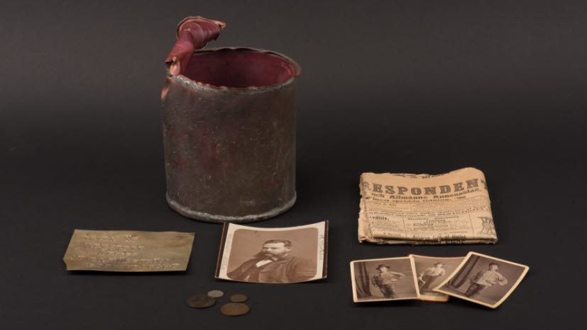 En öppnad burk i metall med fotografier, en graverad metallbit, mynt och tidning placerade framför och vid sidan. 