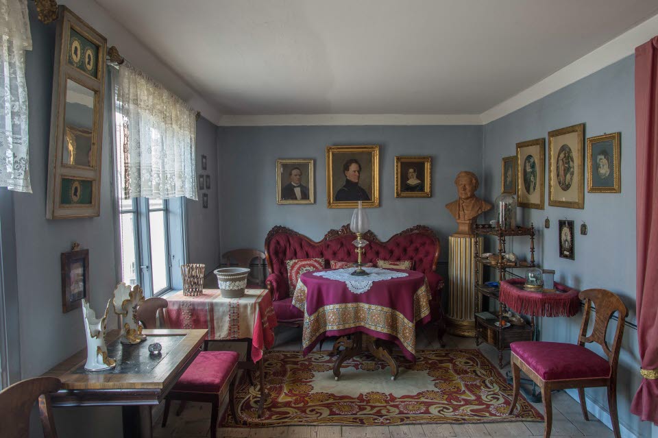 Interiör i museilägenheten i Onkel Adamsgården med möbler klädda i lila tyg, en spegel och ett flertal tavlor på väggarna, fönster med spetsgardiner och en piedestal med byst föreställande  Carl Anton Wetterbergh, pseudonymen Onkel Adam. 