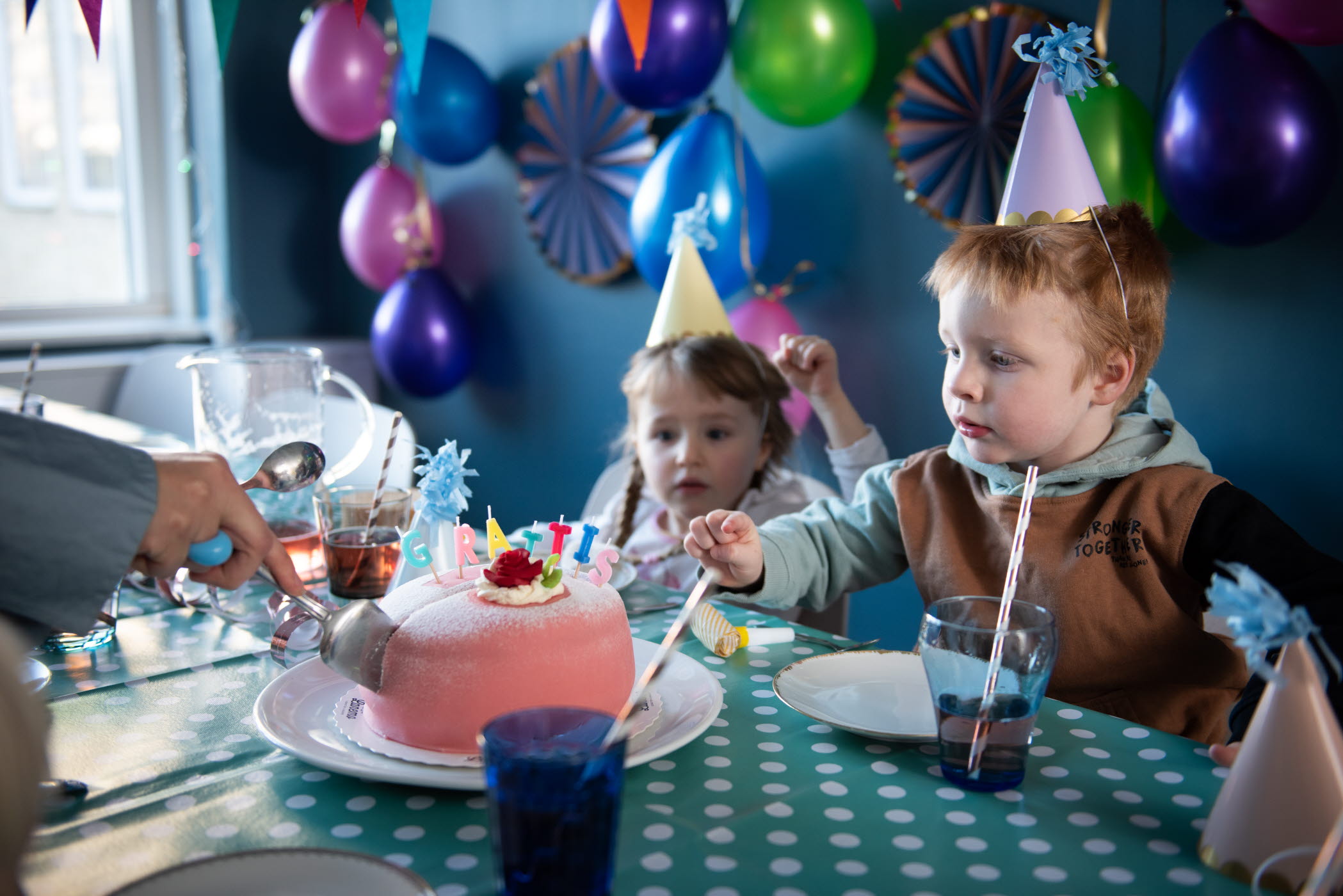 Barn i partyhattar skär upp en tårta som det står "grattis" på. I bakgrunden hänger balonger i olika färger.